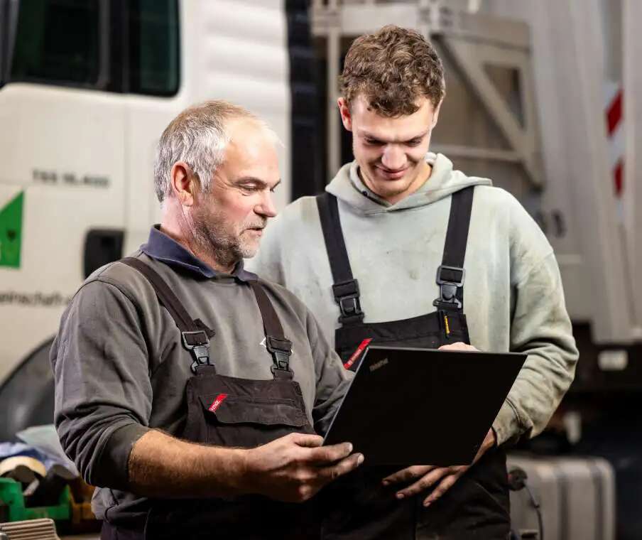 Zwei Männer in Arbeitskleidung stehen vor einem Lkw und schauen gemeinsam auf einen aufgeklappten Laptop. Einer der Männer hält das Gerät in den Händen, während der andere daneben steht. Im Hintergrund ist ein Teil des Lkw zu sehen.