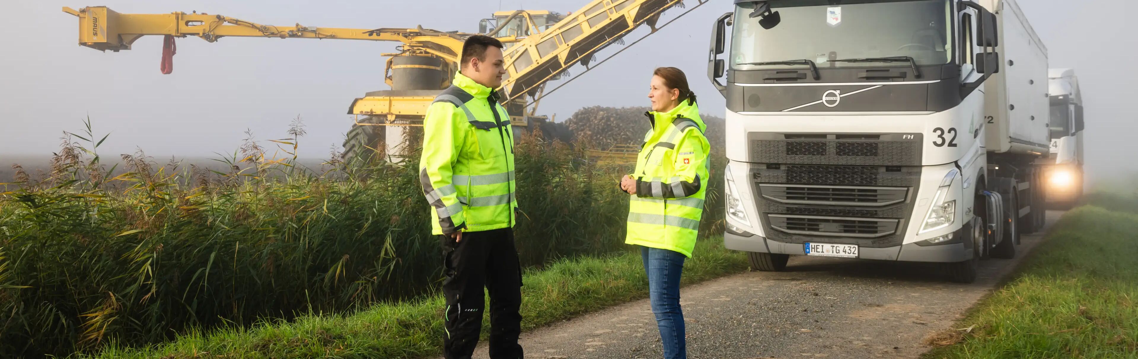 Zwei Personen in gelben Warnjacken stehen auf einem Feldweg vor einem Lkw und sprechen miteinander. Im Hintergrund befindet sich eine gelbe Erntemaschine mit Förderband sowie ein großer Rübenhaufen. Nebel liegt über der Landschaft.