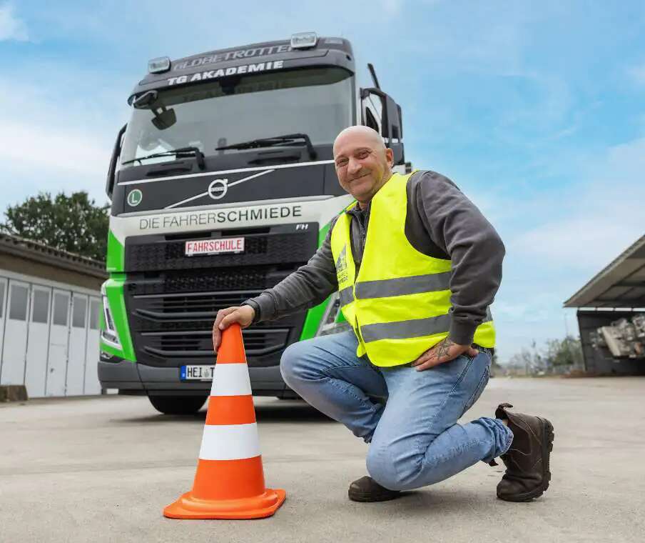 Ein Mann in Warnweste kniet neben einem Verkehrsleitkegel auf einem Hofgelände. Hinter ihm steht ein Lkw mit der Aufschrift „Die Fahrerschmiede“ und einem Schild mit „Fahrschule“. Im Hintergrund sind Gebäude und ein teils bewölkter Himmel zu sehen.