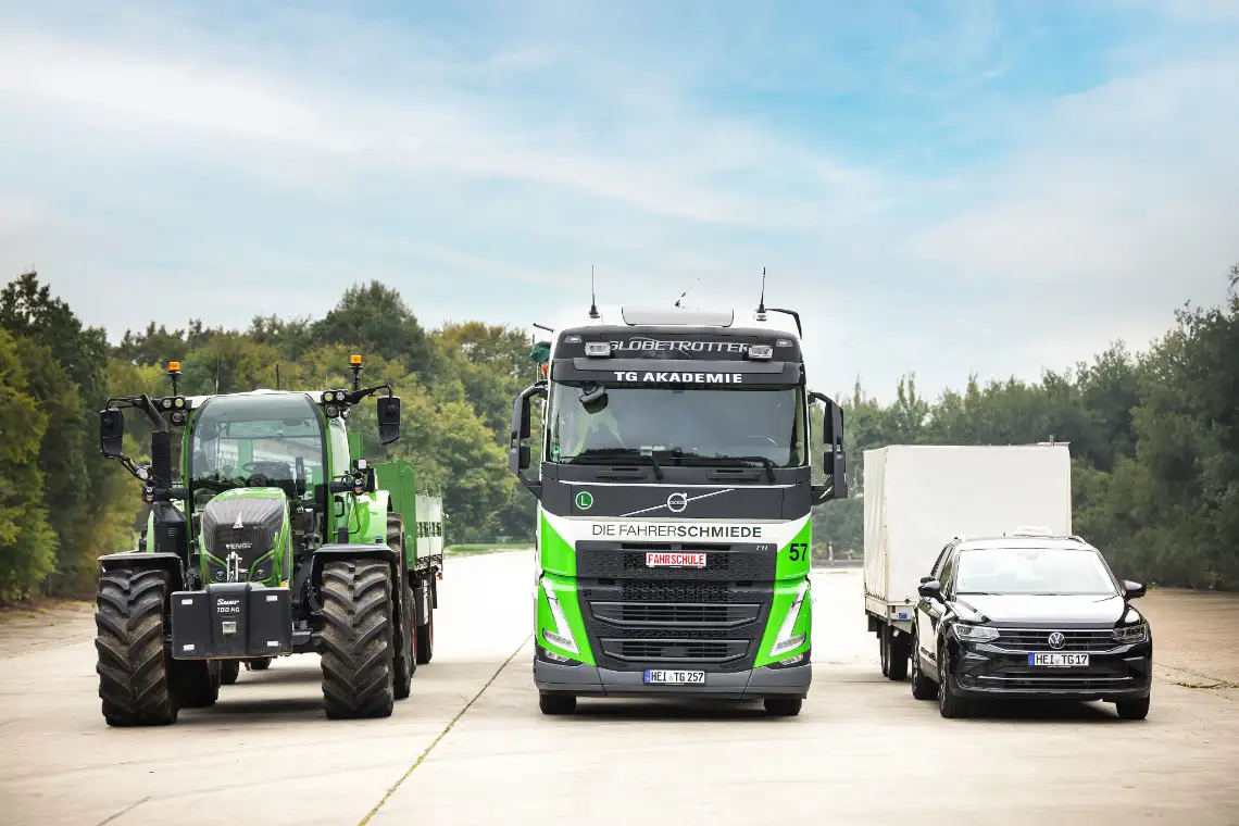 Ein Traktor, ein Lkw und ein Pkw mit Anhänger stehen nebeneinander auf einem betonierten Platz. Die Fahrzeuge gehören zur Fahrerschmiede TG Akademie und sind in Grün, Weiß und Schwarz gestaltet. Im Hintergrund sind Bäume und Himmel zu sehen.